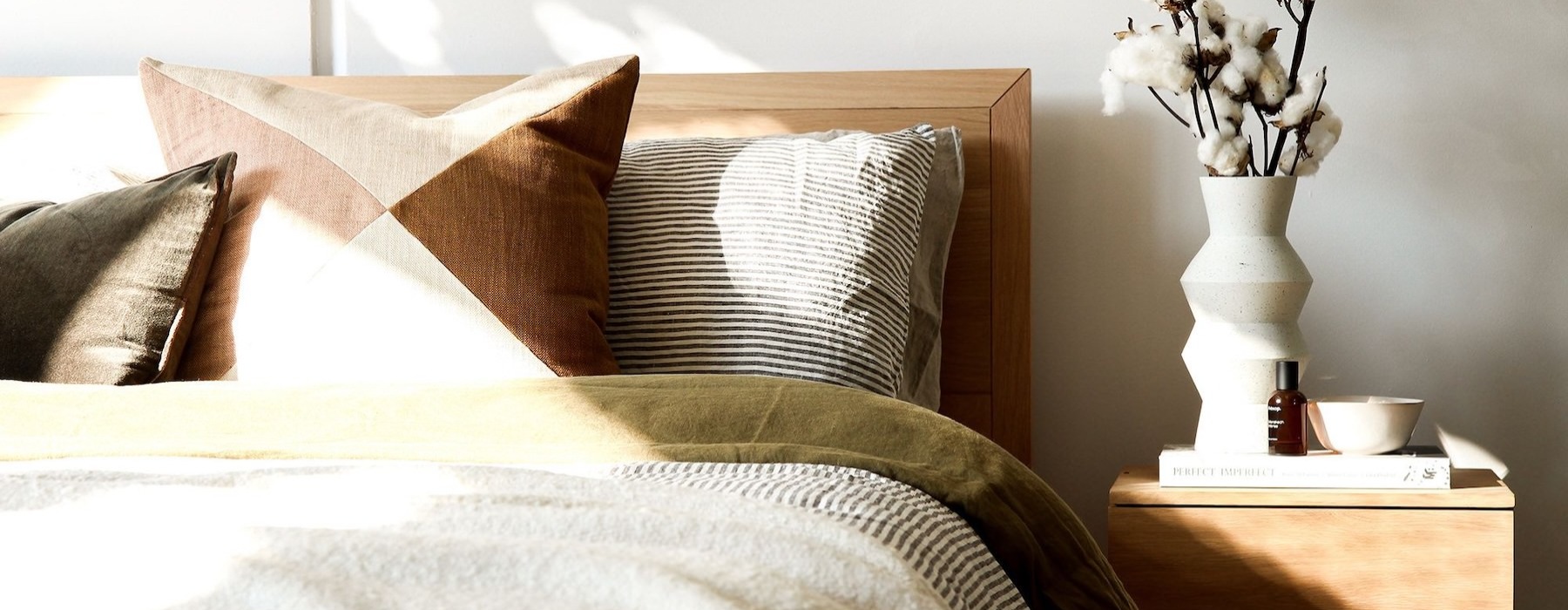 swath of sunlight shines across a well made bed and bedside table with a vase of cotton plants